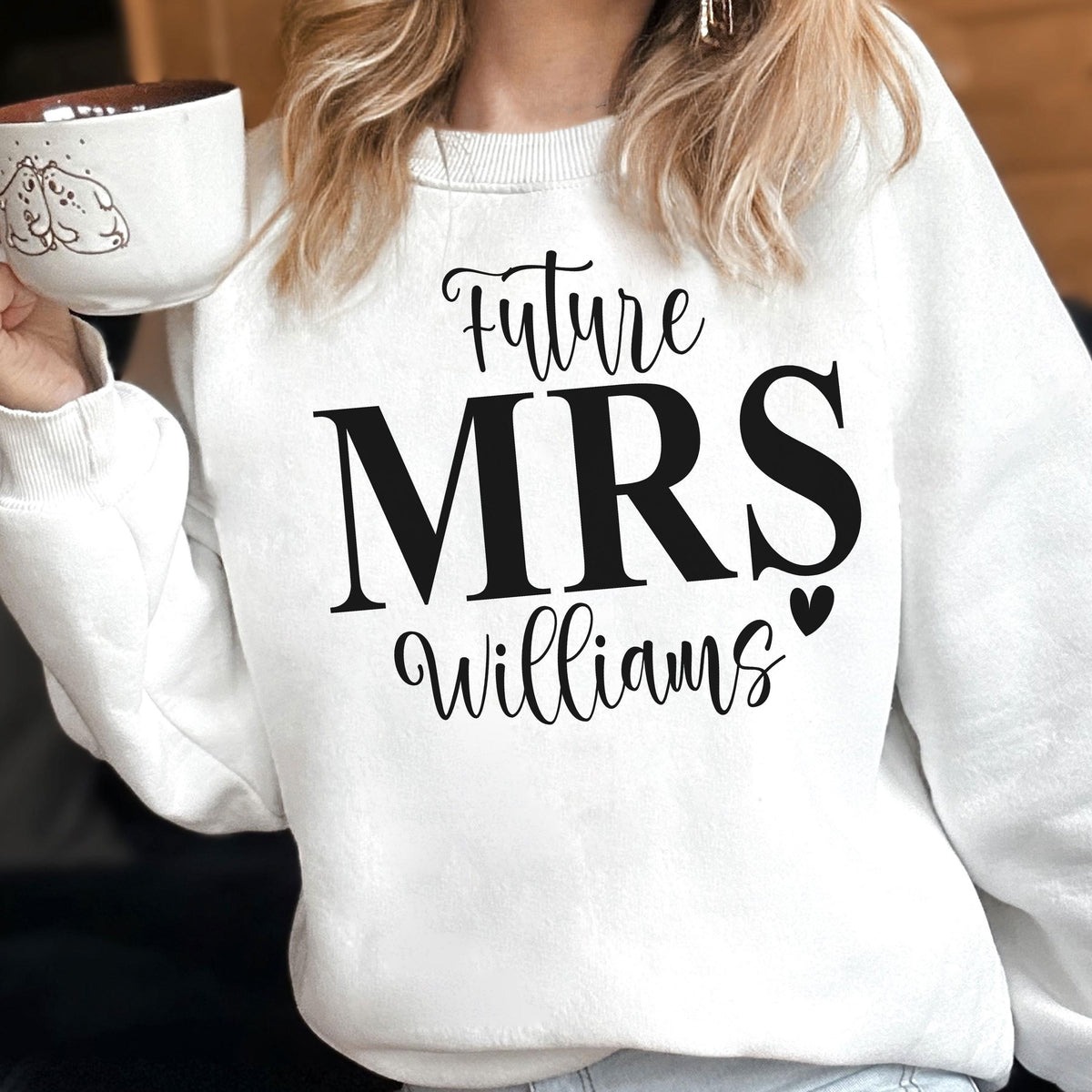 May include: A white long-sleeve crewneck sweatshirt featuring black text that reads Future MRS Williams with a small heart icon. The sweatshirt is made of a soft fabric and is worn over a light-colored garment, paired with a white ceramic mug.