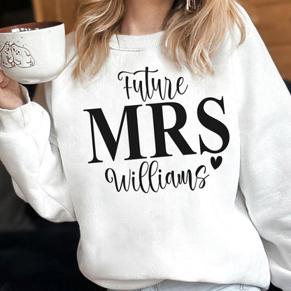 May include: A white long-sleeve crewneck sweatshirt featuring black text that reads Future MRS Williams with a small heart icon. The sweatshirt is made of a soft fabric and is worn over a light-colored garment, paired with a white ceramic mug.