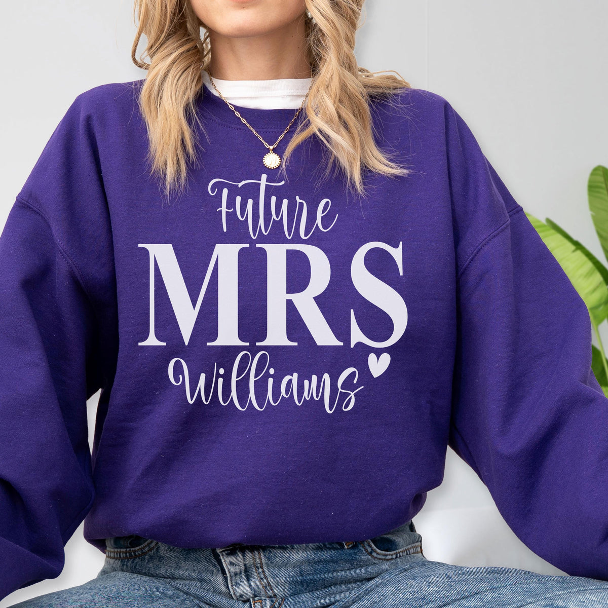 May include: A purple long-sleeve crewneck sweatshirt featuring white text that reads Future MRS Williams with a small heart icon. The garment is worn over a white collared shirt and is paired with blue denim jeans, showcasing a casual style.