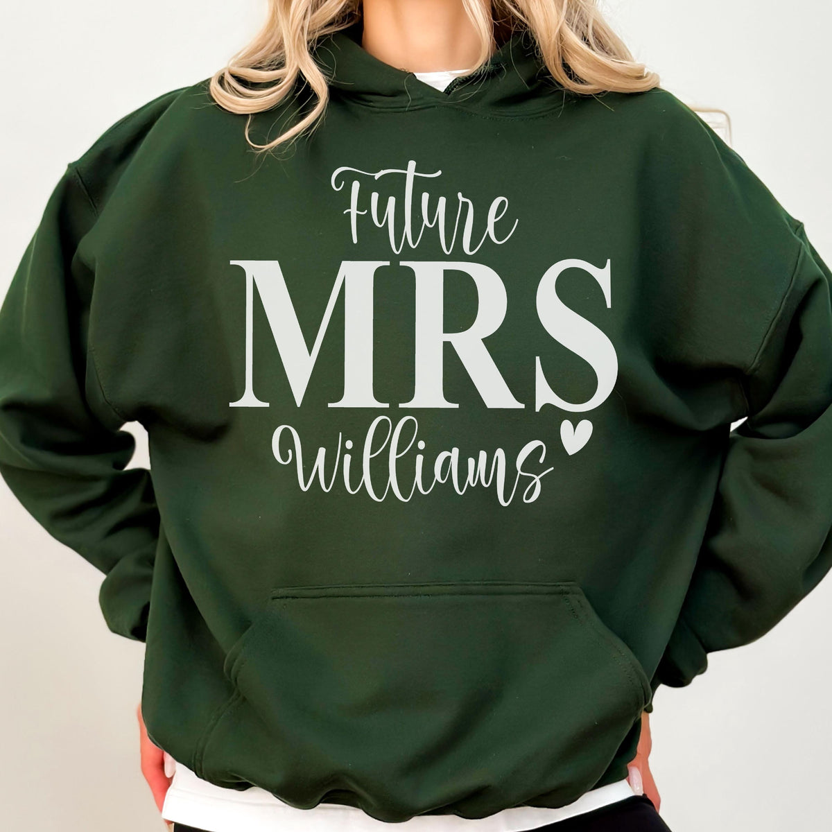 May include: A dark green hooded sweatshirt featuring the white text Future MRS Williams with a small heart icon. The garment includes a front kangaroo pocket, long sleeves, and a hood with matching drawstrings, presented in a standard pullover style.
