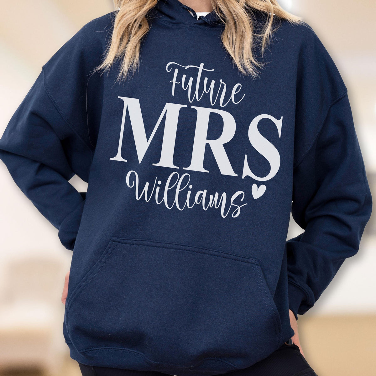 May include: A navy blue hooded sweatshirt featuring white text that reads Future MRS Williams with a small heart icon. The garment includes long sleeves, a hood with drawstrings, and a large front kangaroo pocket for storage and warmth.