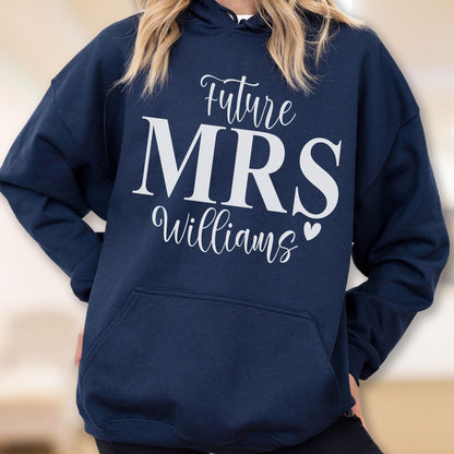 May include: A navy blue hooded sweatshirt featuring white text that reads Future MRS Williams with a small heart icon. The garment includes long sleeves, a hood with drawstrings, and a large front kangaroo pocket for storage and warmth.