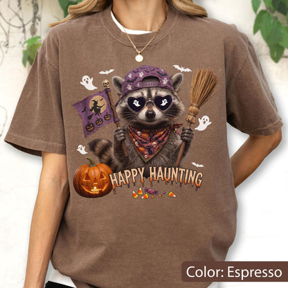 May include: An espresso-colored t-shirt featuring a graphic of a raccoon wearing a purple baseball cap and heart-shaped sunglasses. The raccoon holds a broom and a purple flag with a witch silhouette. Text reads HAPPY HAUNTING with pumpkins.