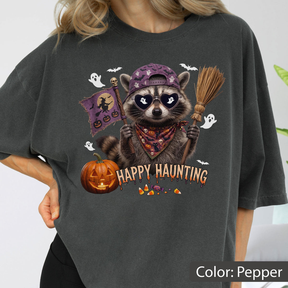 May include: A pepper-colored t-shirt featuring a graphic of a raccoon wearing a purple baseball cap, heart-shaped sunglasses, and a patterned bandana. The raccoon holds a flag and a broom near a jack-o'-lantern, with the text HAPPY HAUNTING.