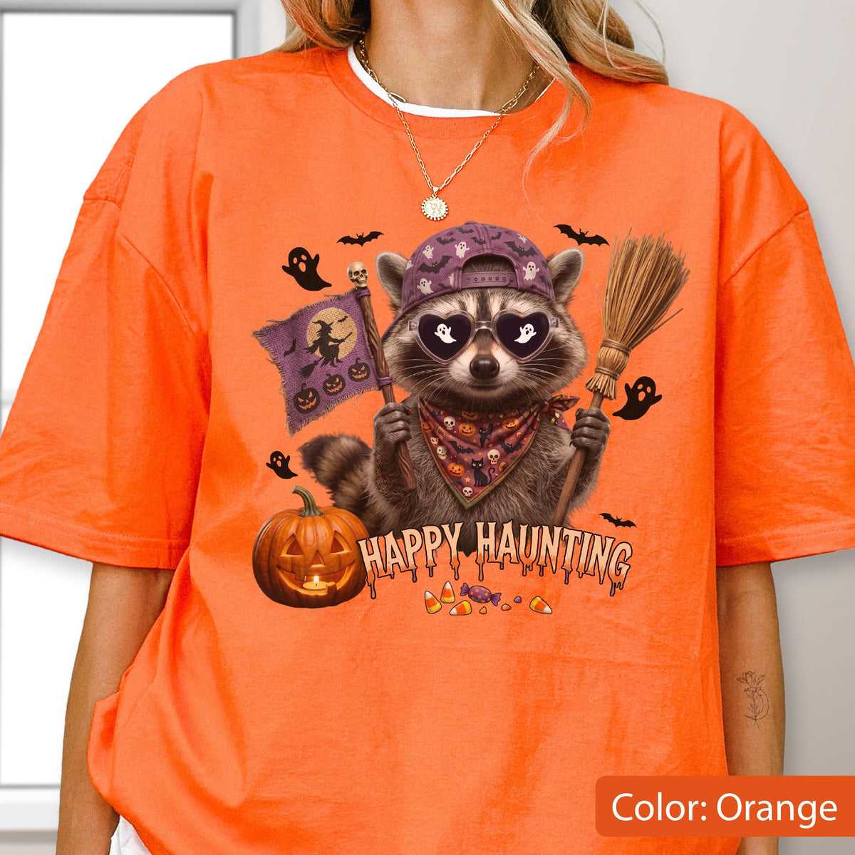 May include: An orange t-shirt featuring a graphic of a raccoon wearing a purple baseball cap and heart-shaped sunglasses. The raccoon holds a broom and a flag with a witch silhouette. Text reads HAPPY HAUNTING above candy corn and a pumpkin.