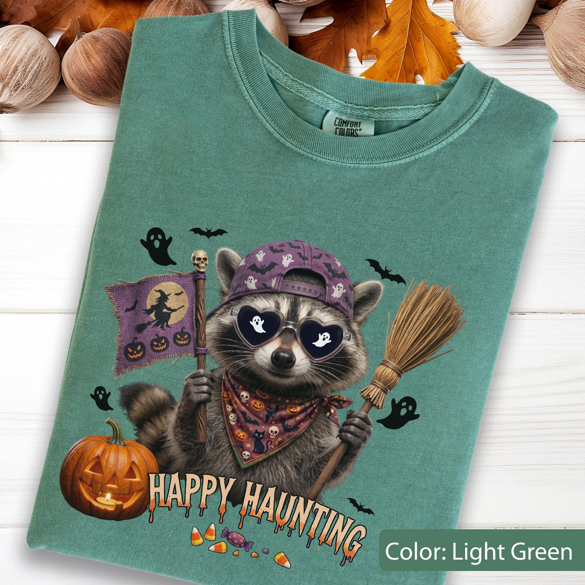 May include: A light green t-shirt featuring a graphic of a raccoon wearing a purple baseball cap and heart-shaped sunglasses. The raccoon holds a flag with a witch silhouette and a broom. Text reads Happy Haunting with pumpkins and ghosts.
