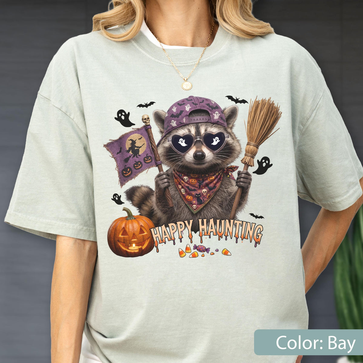 May include: A light green t-shirt featuring a graphic of a raccoon wearing a purple baseball cap and heart-shaped sunglasses. The raccoon holds a broom and a flag with a witch silhouette. Text reads HAPPY HAUNTING above candy corn and a pumpkin.
