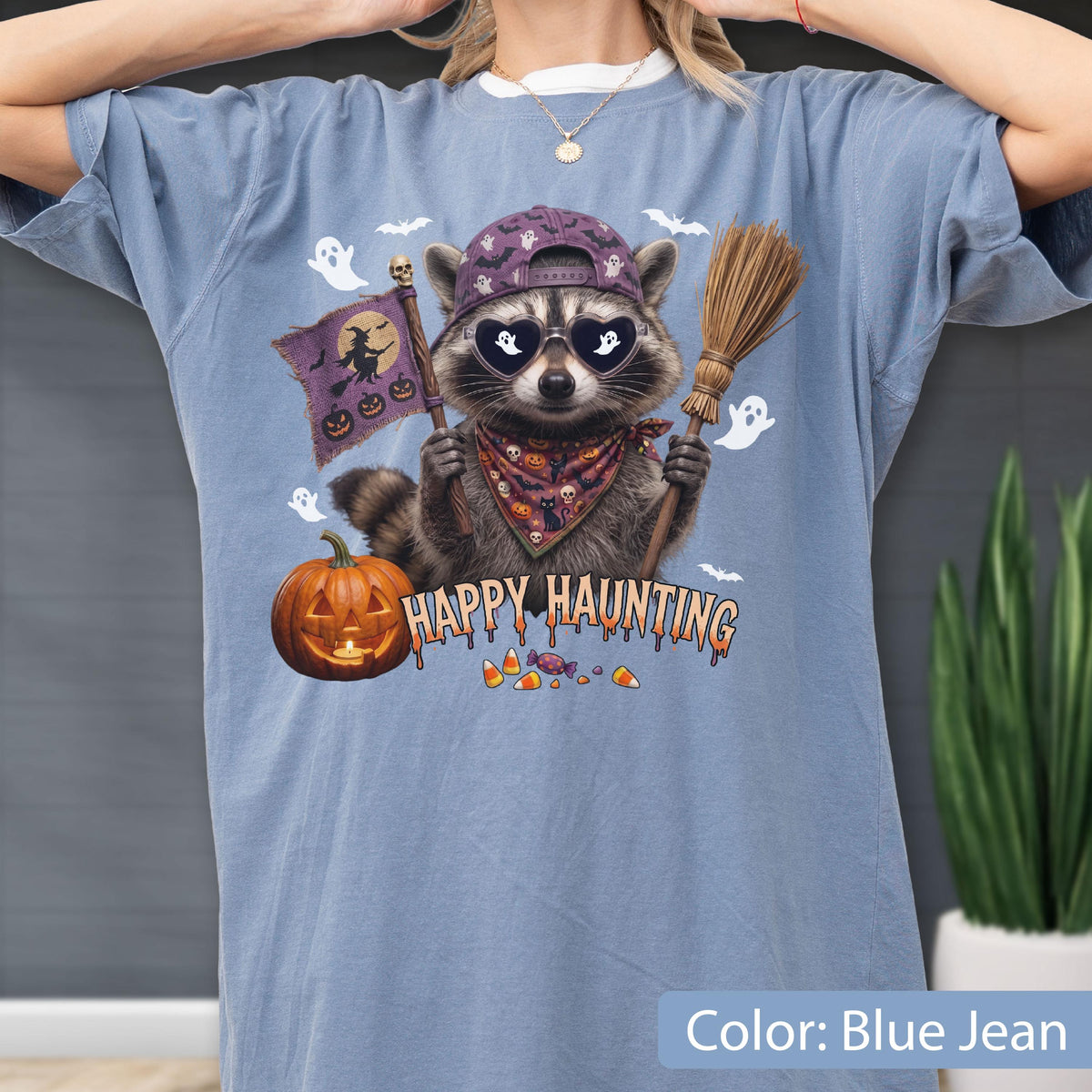May include: A blue jean colored t-shirt featuring a graphic of a raccoon wearing a purple baseball cap, heart-shaped sunglasses with ghost reflections, and a patterned bandana. The raccoon holds a broom and a flag with the text Happy Haunting.