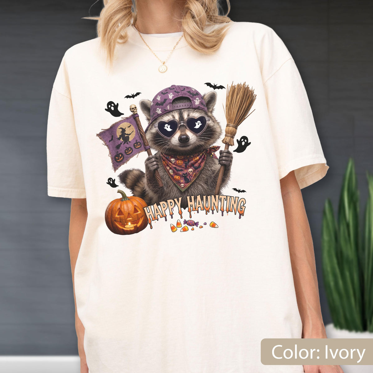 May include: An ivory t-shirt featuring a graphic of a raccoon wearing a purple baseball cap and heart-shaped sunglasses. The raccoon holds a broom and a flag with a witch silhouette. Text reads Happy Haunting, with a pumpkin, ghosts, and candy.