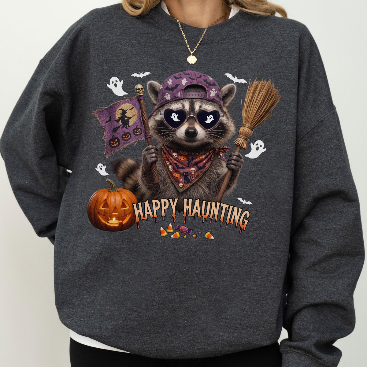 May include: A dark gray sweatshirt featuring a graphic of a raccoon wearing a purple baseball cap, heart-shaped sunglasses, and a patterned bandana. The raccoon holds a broom and a purple flag with a witch silhouette. Text reads HAPPY HAUNTING.
