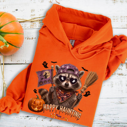 May include: An orange hooded sweatshirt featuring a graphic of a raccoon wearing a purple baseball cap and heart-shaped sunglasses. The raccoon holds a broom and a flag with a witch silhouette. Text reads Happy Haunting with pumpkins.