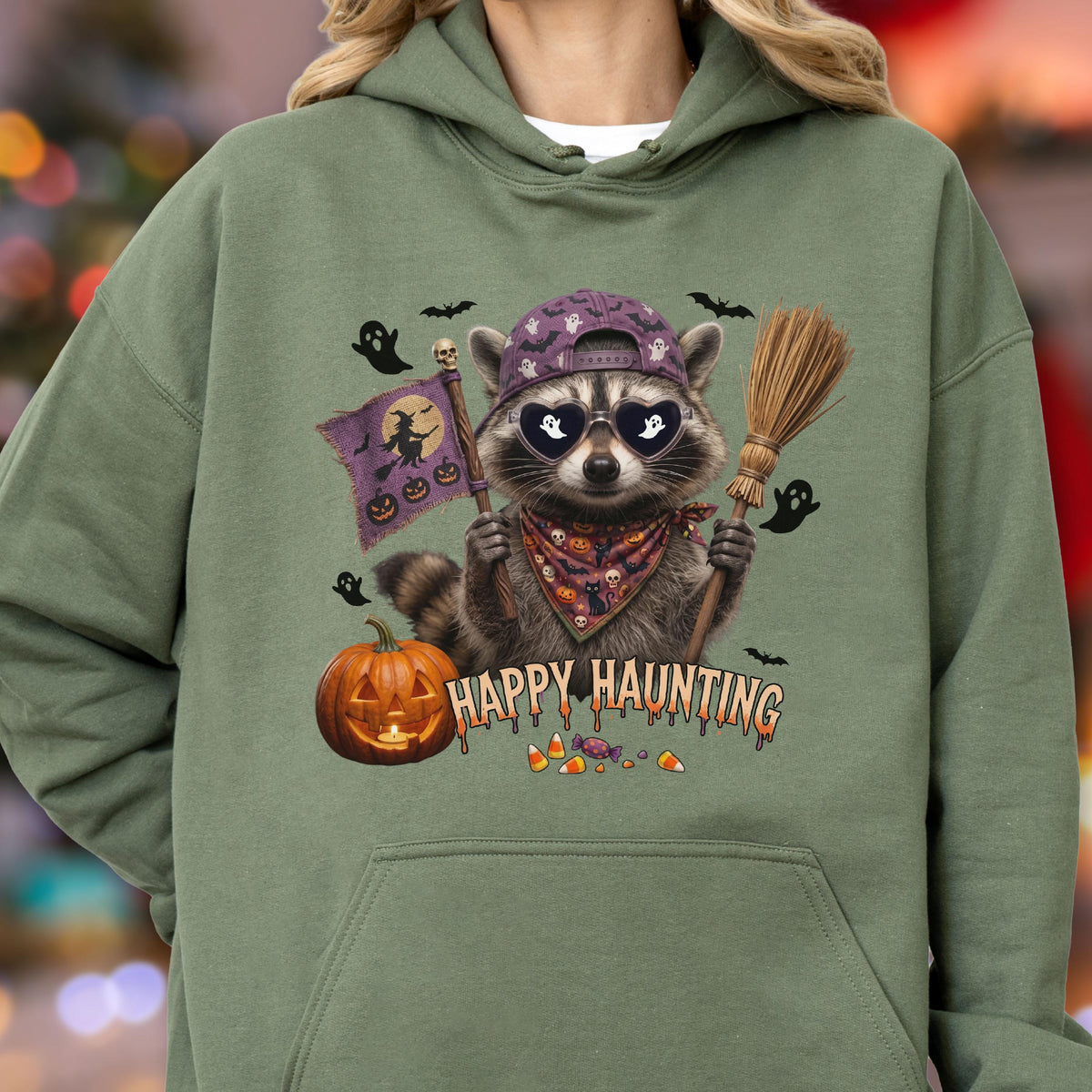 May include: An olive green hooded sweatshirt featuring a graphic of a raccoon wearing a purple baseball cap and heart-shaped sunglasses. The raccoon holds a broom and a flag with a witch silhouette. Text reads HAPPY HAUNTING with pumpkins and candy.