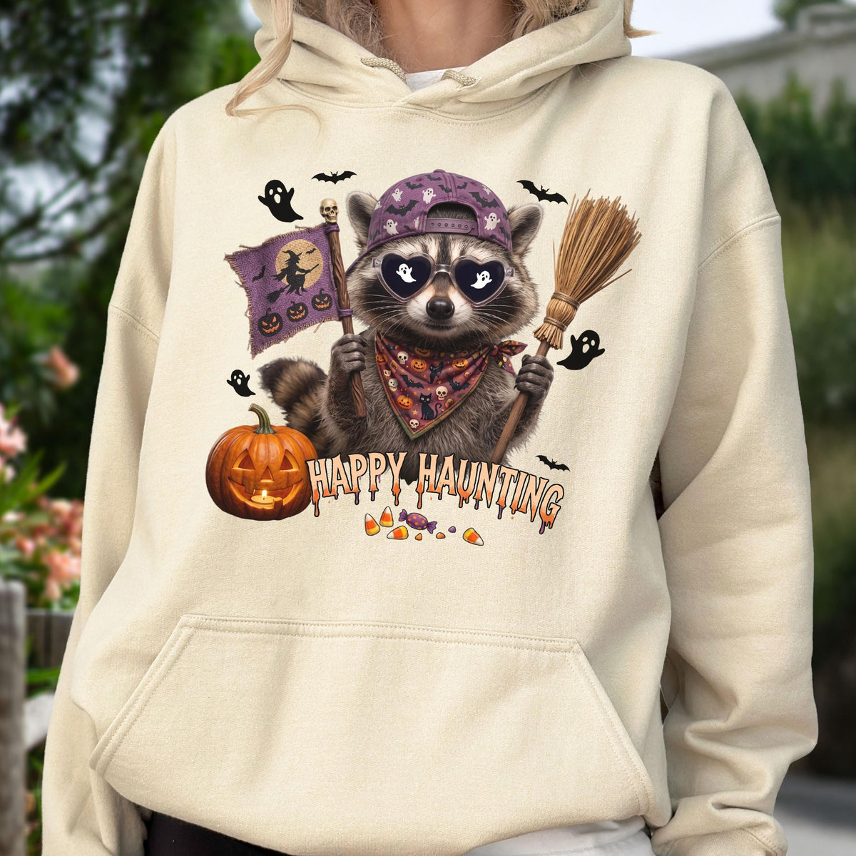 May include: A cream-colored hooded sweatshirt featuring a graphic of a raccoon wearing a purple baseball cap, heart-shaped sunglasses with ghost silhouettes, and a patterned bandana. The raccoon holds a flag and broom, with text reading HAPPY HAUNTING.