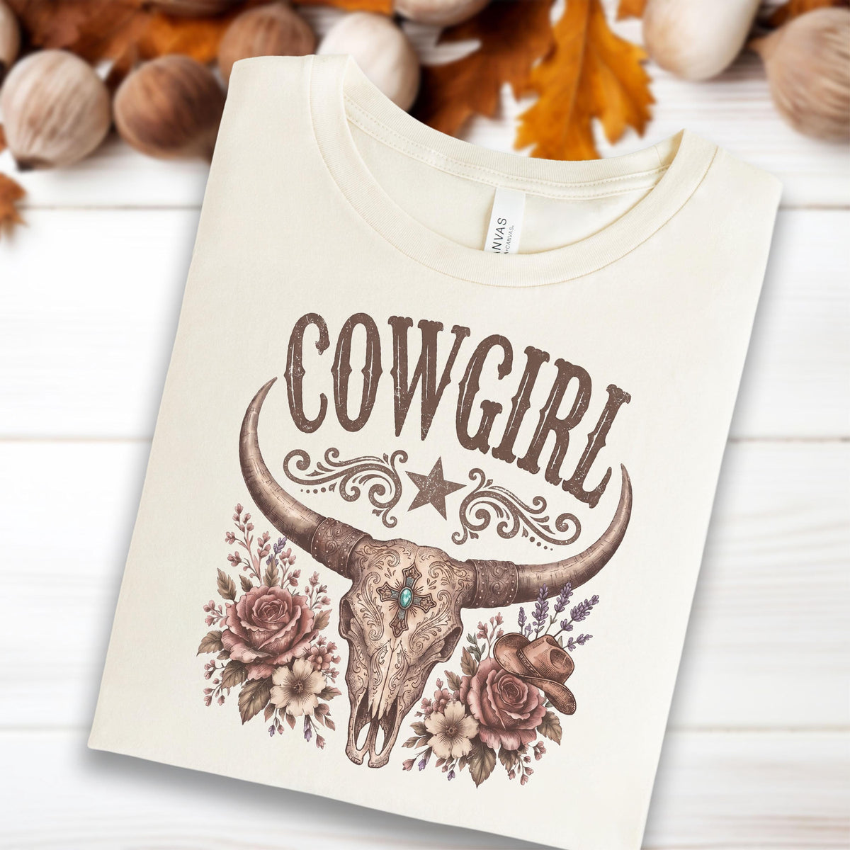 May include: A cream-colored t-shirt featuring a brown graphic print of a cow skull with long horns, a central cross, and floral accents. The word COWGIRL is printed in a decorative brown font above the skull, accompanied by a small star and swirls.