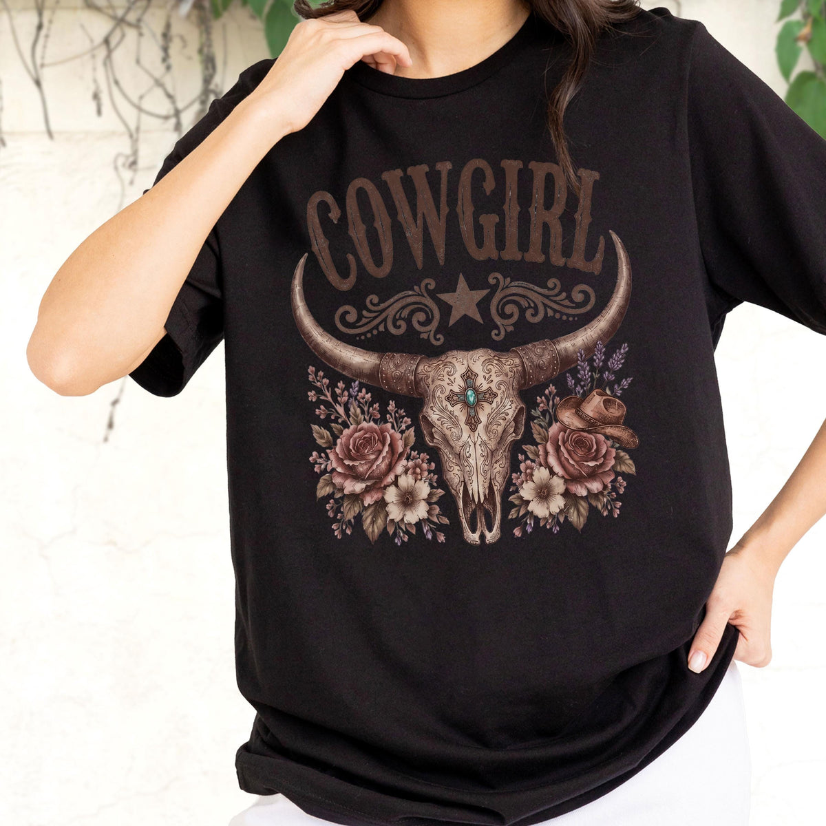 May include: A black short-sleeved t-shirt featuring a graphic print of a steer skull with a central turquoise stone, surrounded by brown roses and a small hat. The word COWGIRL is printed in a brown western-style font above the skull design.
