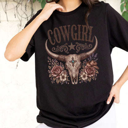 May include: A black short-sleeved t-shirt featuring a graphic print of a steer skull with a central turquoise stone, surrounded by brown roses and a small hat. The word COWGIRL is printed in a brown western-style font above the skull design.