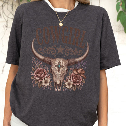 May include: A dark charcoal gray t-shirt featuring a graphic print of a steer skull with a turquoise stone accent, surrounded by roses and a small cowboy hat. The word COWGIRL is printed in a brown, distressed font above the skull illustration.
