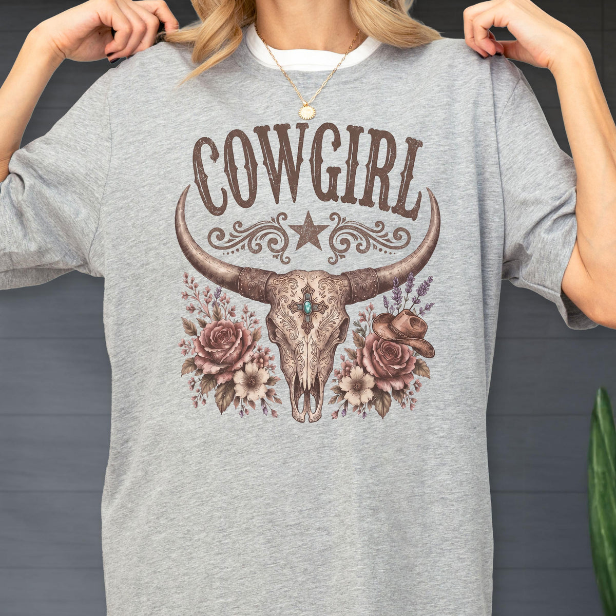 May include: A heather gray t-shirt featuring a graphic print of a longhorn steer skull centered below the text COWGIRL. The design includes a star, floral accents, and a small cowboy hat, all rendered in shades of brown, tan, and muted teal.