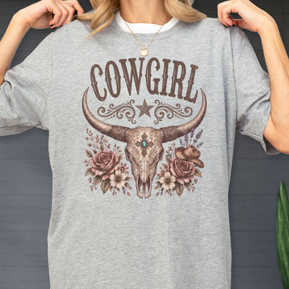 May include: A heather gray t-shirt featuring a graphic print of a longhorn steer skull centered below the text COWGIRL. The design includes a star, floral accents, and a small cowboy hat, all rendered in shades of brown, tan, and muted teal.