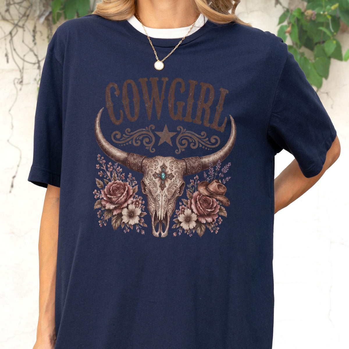 May include: A navy blue t-shirt featuring a graphic print of a longhorn skull with a central turquoise stone, surrounded by brown roses and a small cowboy hat. The word COWGIRL is printed in large, distressed brown letters above the skull.