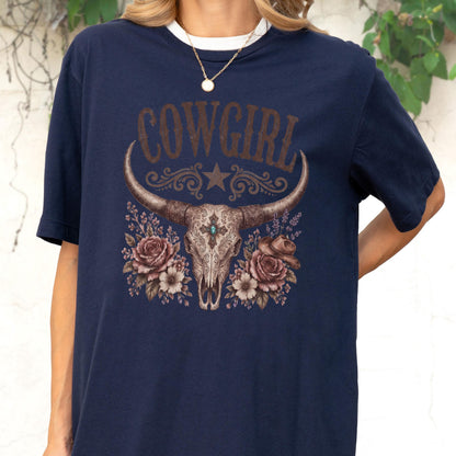 May include: A navy blue t-shirt featuring a graphic print of a longhorn skull with a central turquoise stone, surrounded by brown roses and a small cowboy hat. The word COWGIRL is printed in large, distressed brown letters above the skull.