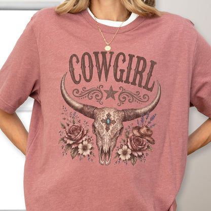 May include: A dusty rose short-sleeve t-shirt featuring a graphic print of a longhorn skull, roses, and a cowboy hat. The text COWGIRL is printed in a dark brown font above the skull, which is decorated with a star and floral accents.