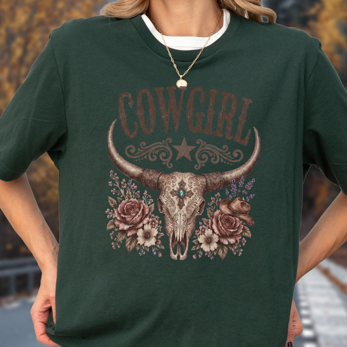 May include: A dark green short-sleeve t-shirt featuring a graphic print of a steer skull with ornate patterns, a central turquoise stone, roses, and a small cowboy hat. The text COWGIRL is printed in a brown distressed font above the skull.