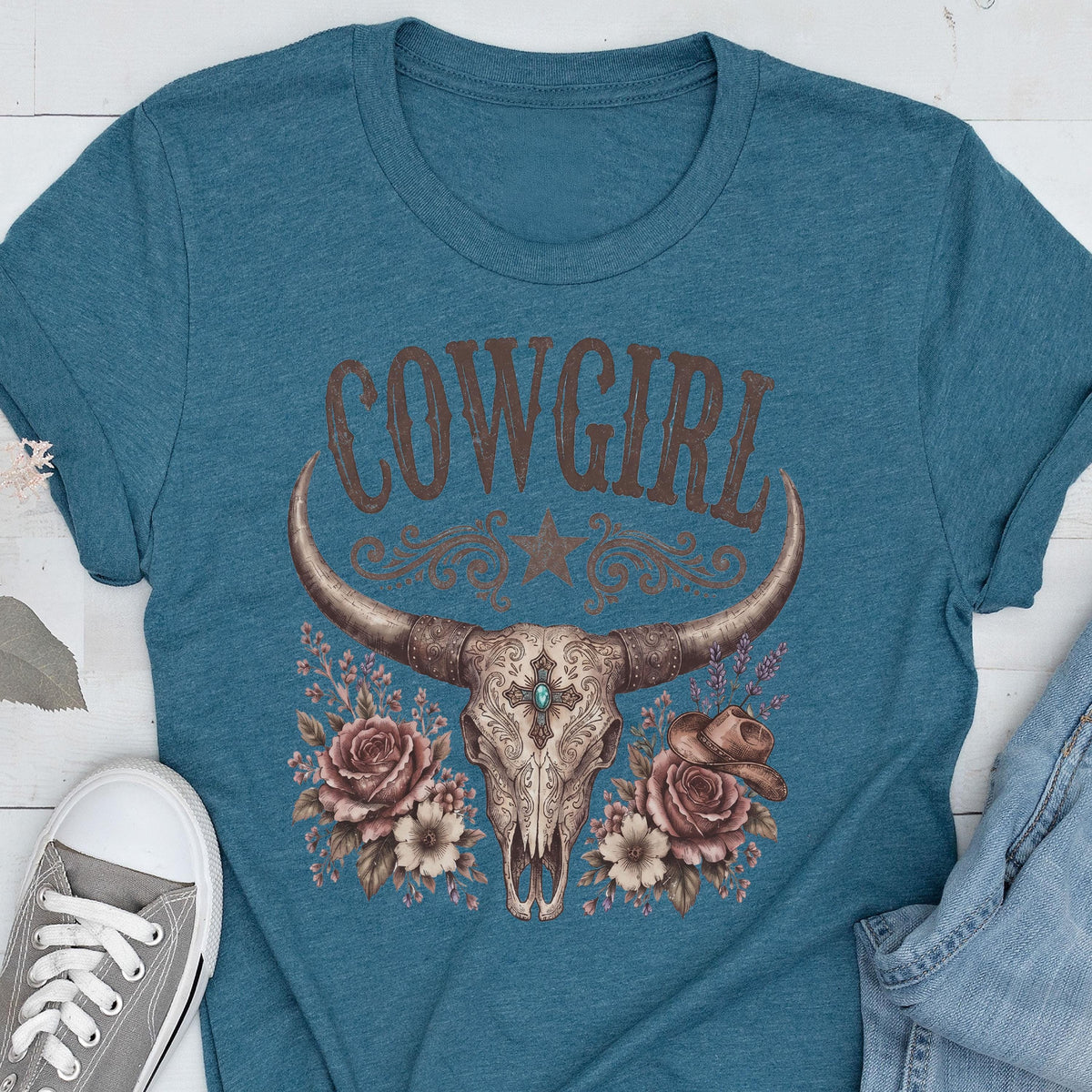 May include: A teal short-sleeved t-shirt featuring a graphic print of a longhorn skull with a central turquoise stone, surrounded by roses and a small cowboy hat. The word COWGIRL is printed in large brown letters above the skull illustration.