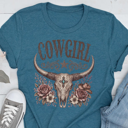 May include: A teal short-sleeved t-shirt featuring a graphic print of a longhorn skull with a central turquoise stone, surrounded by roses and a small cowboy hat. The word COWGIRL is printed in large brown letters above the skull illustration.