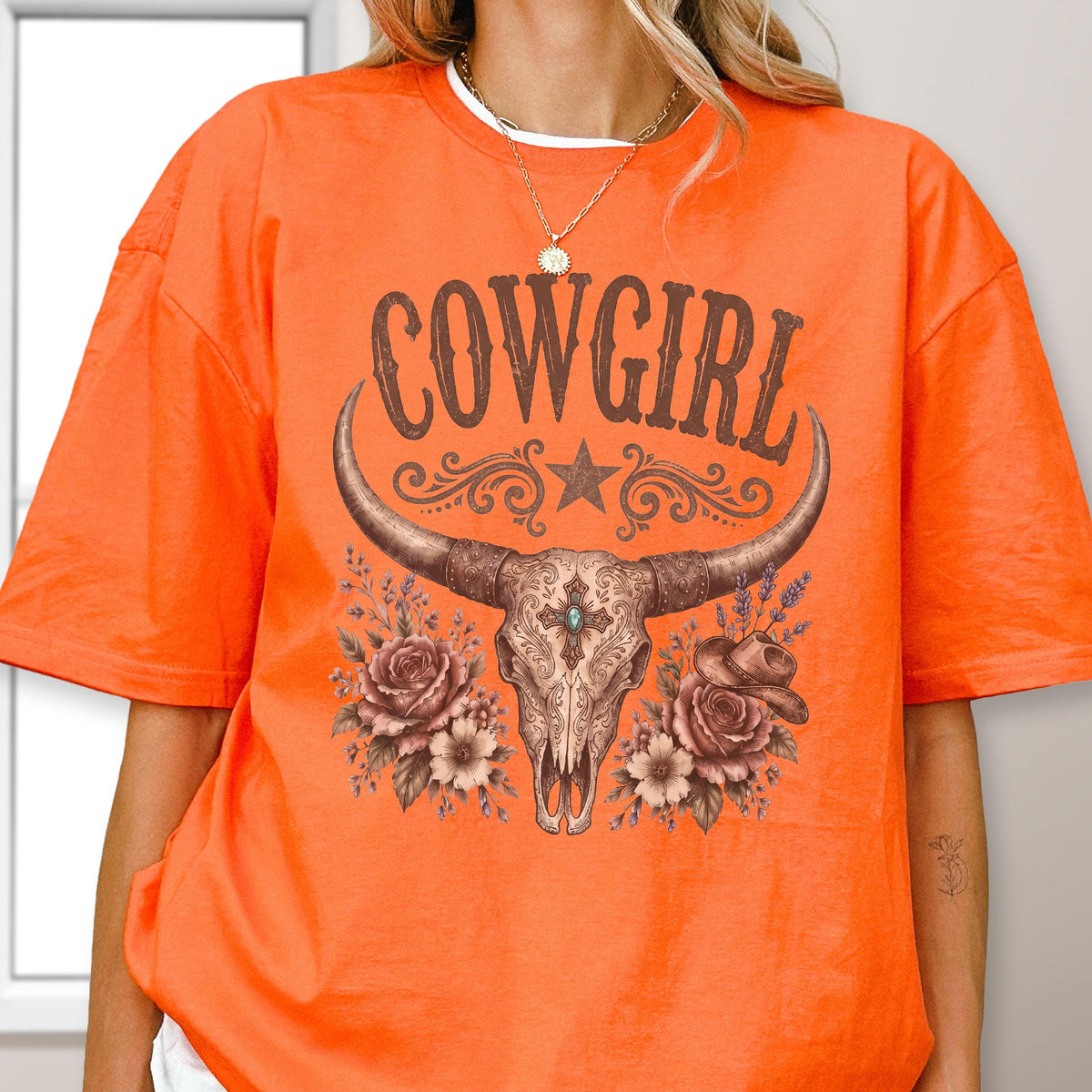 May include: An orange short-sleeved t-shirt featuring a graphic print of a steer skull with long horns, surrounded by roses and a small hat. The text COWGIRL is printed in brown above the skull, which includes a central turquoise accent.