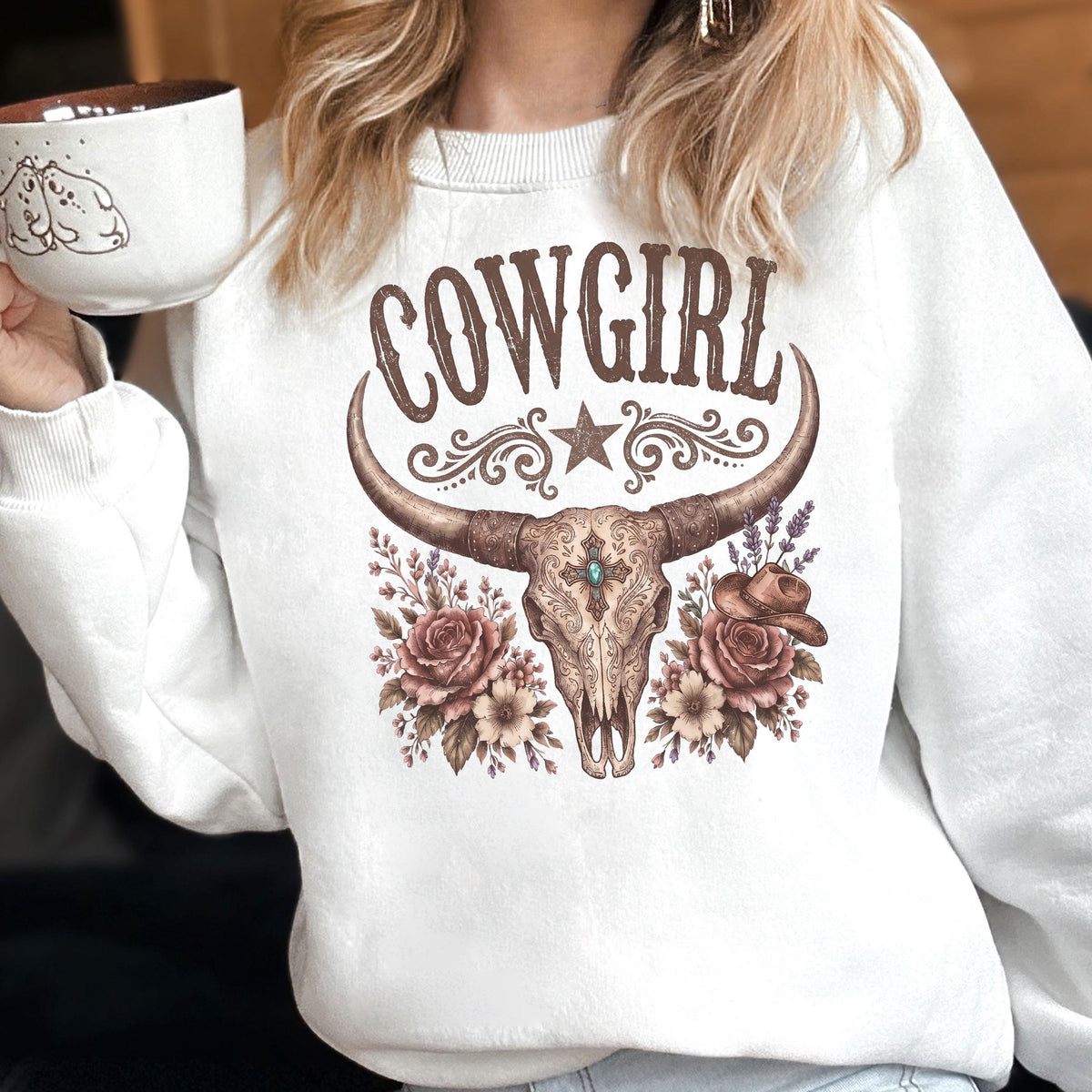 May include: A white crewneck sweatshirt featuring a brown graphic print of a longhorn skull adorned with roses, a small cowboy hat, and the word COWGIRL in a decorative font. The design includes a central star and intricate floral flourishes.