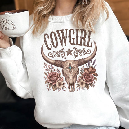 May include: A white crewneck sweatshirt featuring a brown graphic print of a longhorn skull adorned with roses, a small cowboy hat, and the word COWGIRL in a decorative font. The design includes a central star and intricate floral flourishes.