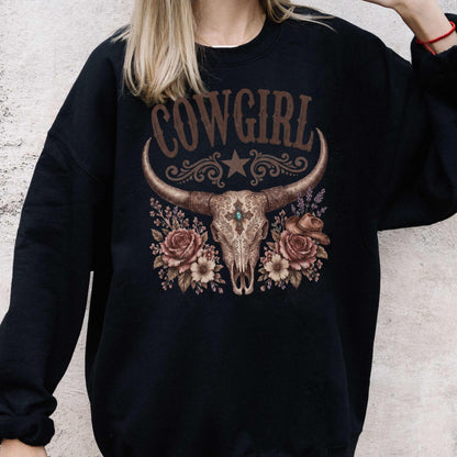 May include: A black crewneck sweatshirt featuring a graphic print of a steer skull with long horns, centered above a cluster of brown roses and a small hat. The word COWGIRL is printed in large brown letters above the skull, with a star and scrollwork.