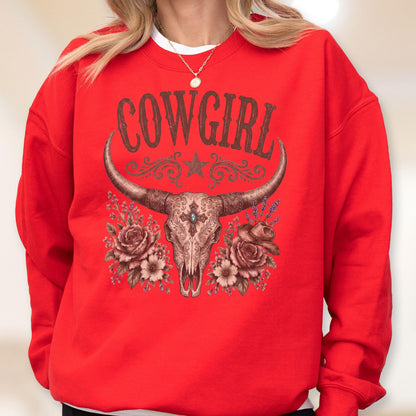 May include: A bright red crewneck sweatshirt featuring a graphic print of a steer skull with long horns, centered above floral arrangements and a small hat. The word COWGIRL is printed in a dark brown, stylized font above the steer skull graphic.