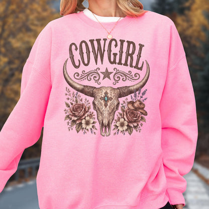 May include: A bright pink long-sleeve sweatshirt featuring a graphic print of a steer skull with long horns, a central star, floral accents, and a small hat. The text COWGIRL is printed in a dark brown font above the skull illustration.