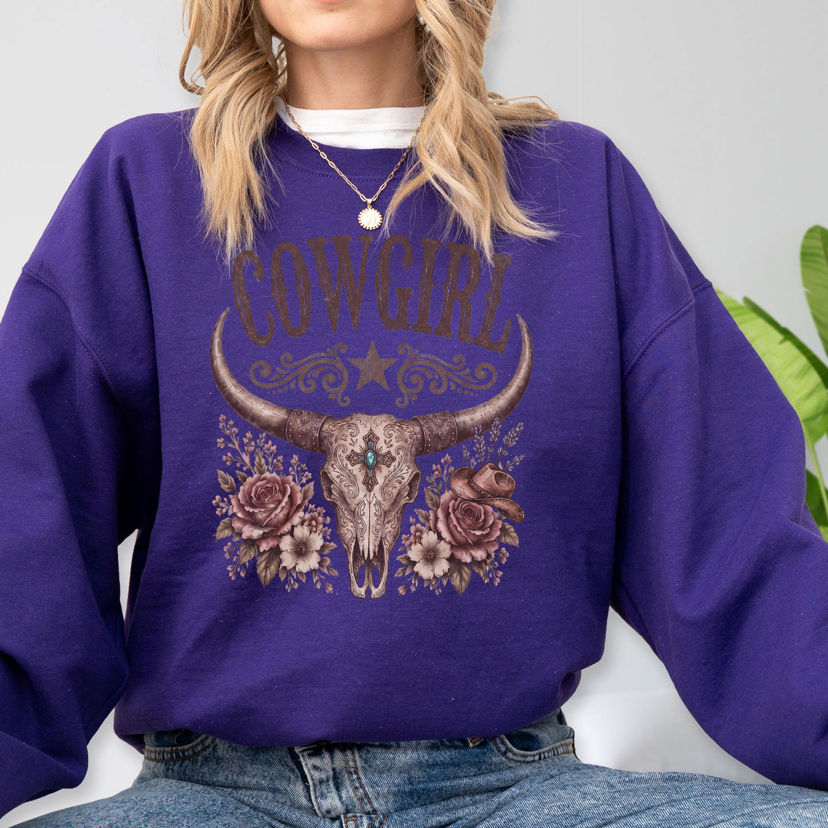 May include: A deep purple long-sleeve sweatshirt featuring a graphic print of a steer skull with long horns, surrounded by floral accents and a small hat. The text COW GIRL is printed in a distressed brown font above the skull illustration.