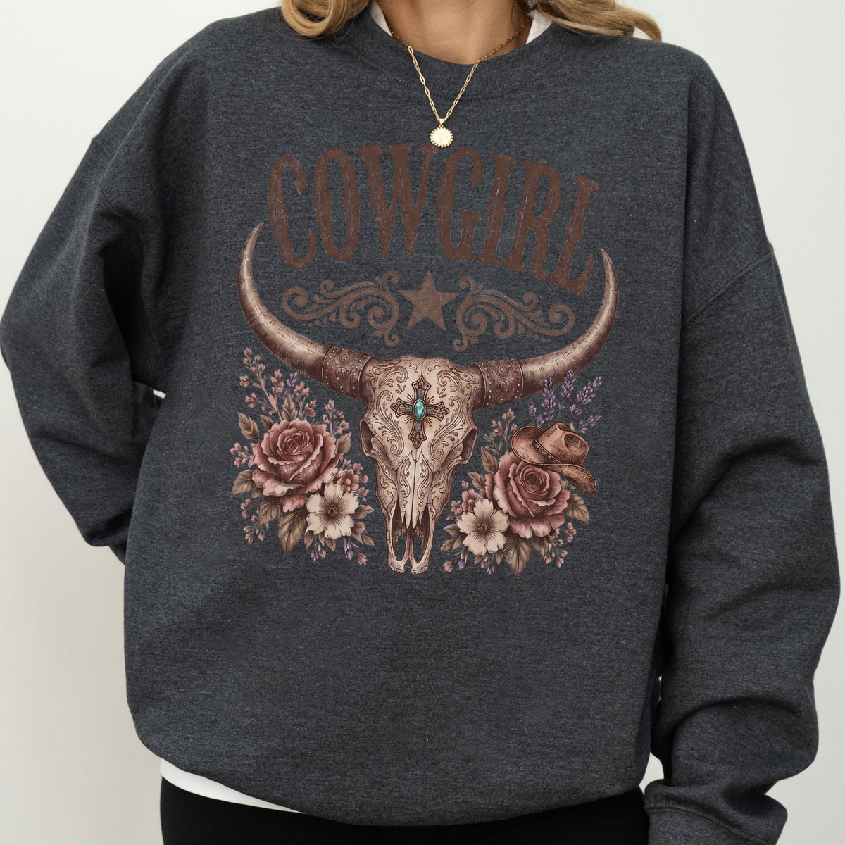 May include: A dark charcoal gray crewneck sweatshirt featuring a graphic print of a longhorn steer skull centered on the chest. The skull is adorned with a turquoise stone, surrounded by brown roses, a small cowboy hat, and the text COWGIRL.