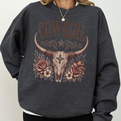 May include: A dark charcoal gray crewneck sweatshirt featuring a graphic print of a longhorn steer skull centered on the chest. The skull is adorned with a turquoise stone, surrounded by brown roses, a small cowboy hat, and the text COWGIRL.