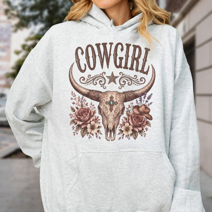 May include: A light gray hooded sweatshirt featuring a graphic print of a steer skull with long horns, a central turquoise stone, and floral accents. The text COWGIRL is printed in brown above the skull, accompanied by a star and scrollwork.