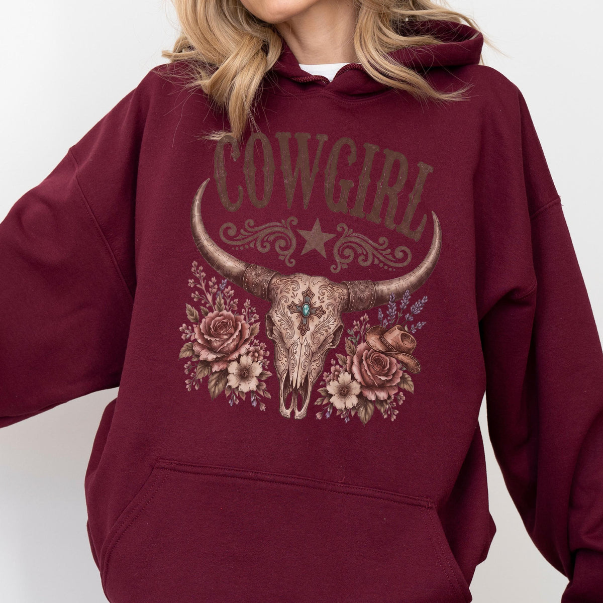 May include: A maroon hooded sweatshirt featuring a graphic print of a longhorn skull adorned with roses, a small cowboy hat, and a central turquoise stone. The text COWGIRL is printed in a distressed font above the skull, with a star and scrollwork.