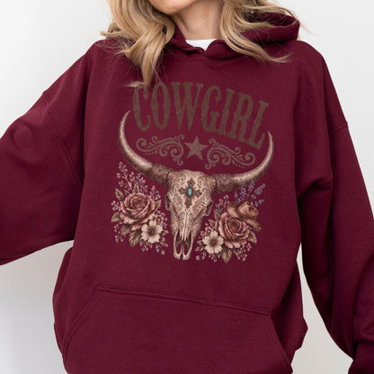 May include: A maroon hooded sweatshirt featuring a graphic print of a longhorn skull adorned with roses, a small cowboy hat, and a central turquoise stone. The text COWGIRL is printed in a distressed font above the skull, with a star and scrollwork.