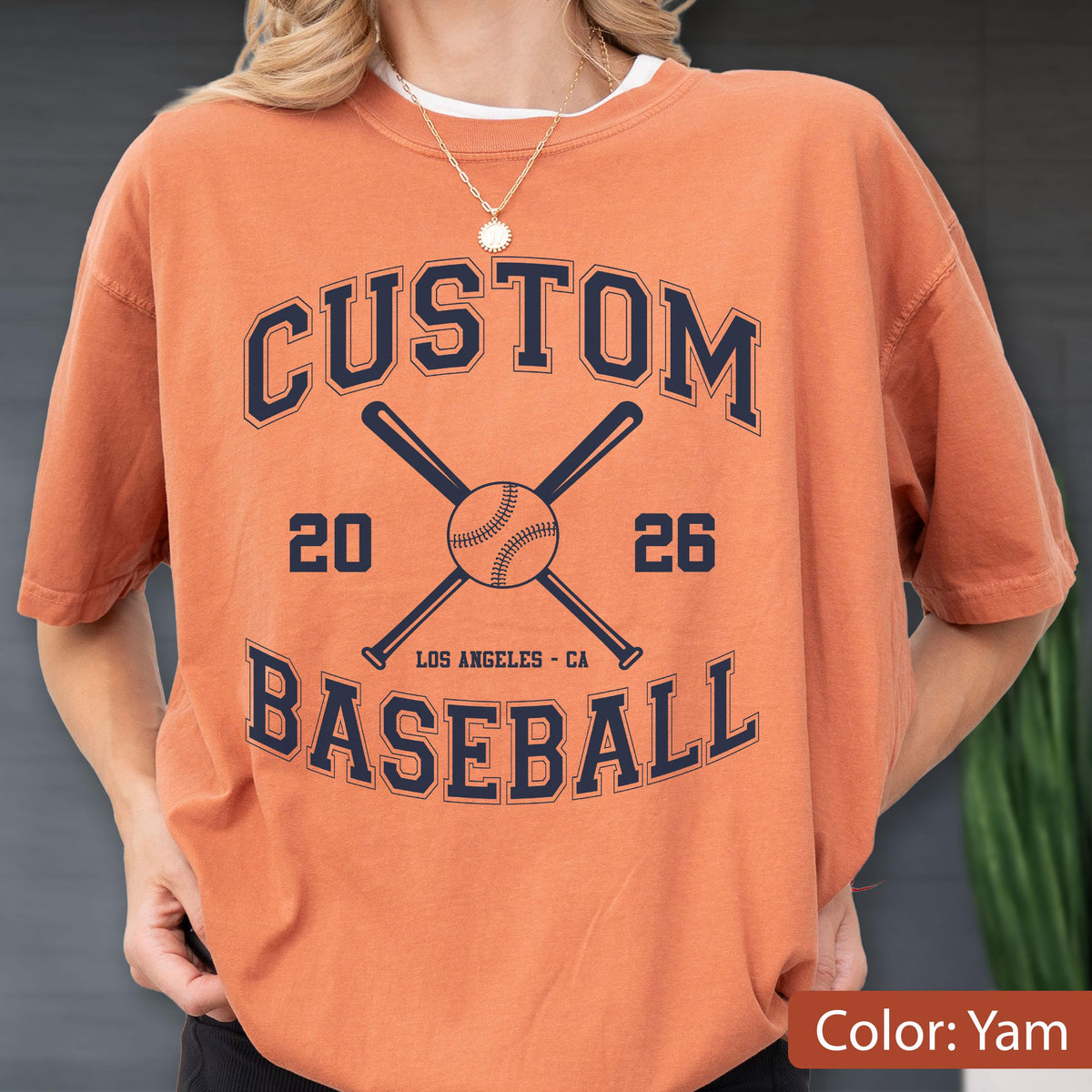 May include: A yam-colored short-sleeve t-shirt featuring a dark navy blue graphic print. The design includes the text CUSTOM BASEBALL, the numbers 20 and 26, and an illustration of two crossed baseball bats behind a central baseball.