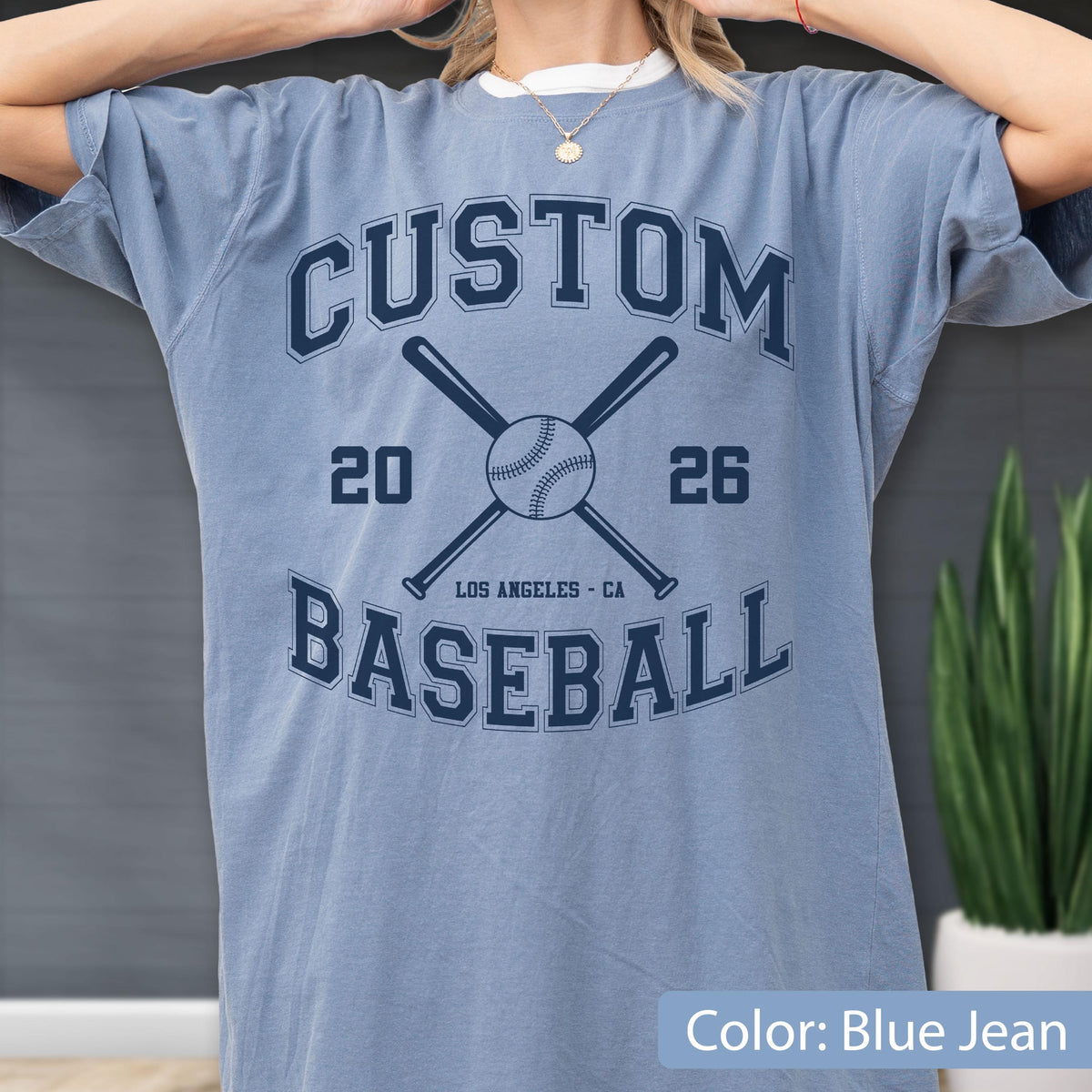 May include: A blue jean colored t-shirt featuring a dark navy graphic with the text CUSTOM BASEBALL, the numbers 20 and 26, and an illustration of a baseball with two crossed bats. The text LOS ANGELES - CA is printed below the baseball.