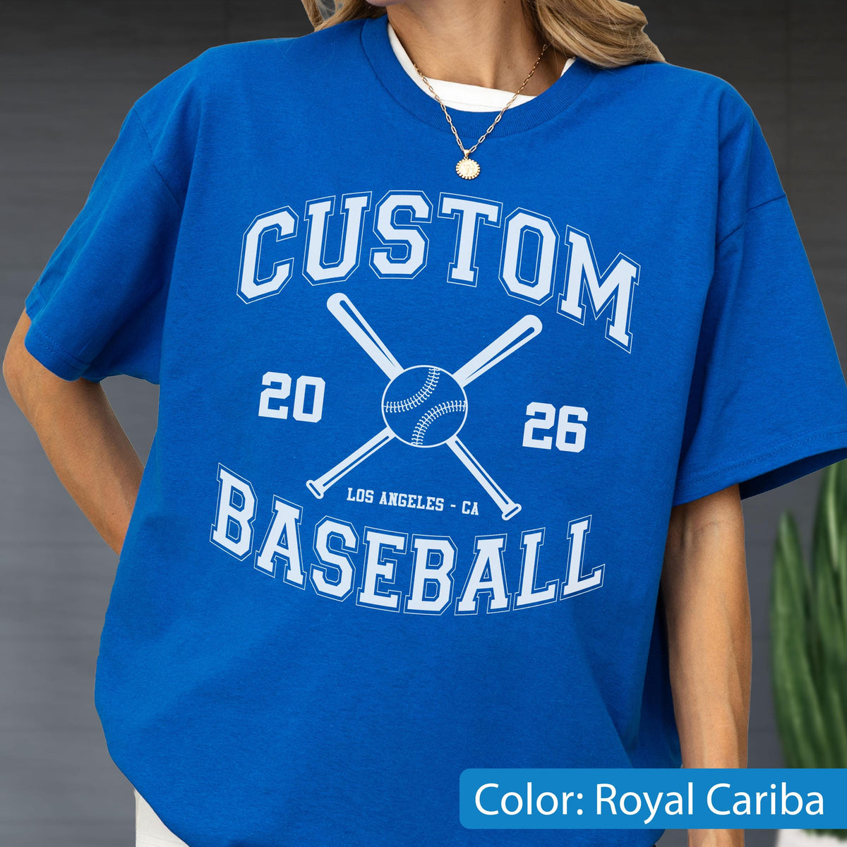 May include: A royal blue short-sleeve t-shirt featuring white screen-printed text that reads CUSTOM BASEBALL with the numbers 20 and 26 on either side of a central graphic showing two crossed baseball bats and a baseball, with LOS ANGELES - CA below.