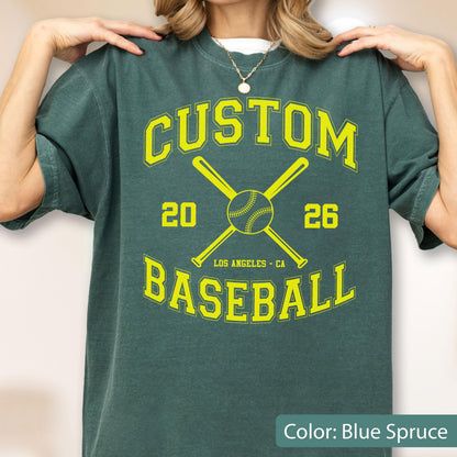 May include: A blue spruce t-shirt featuring a yellow graphic with the text CUSTOM BASEBALL 20 26 LOS ANGELES CA. The design includes two crossed baseball bats and a baseball in the center, printed in a bold, collegiate-style font.