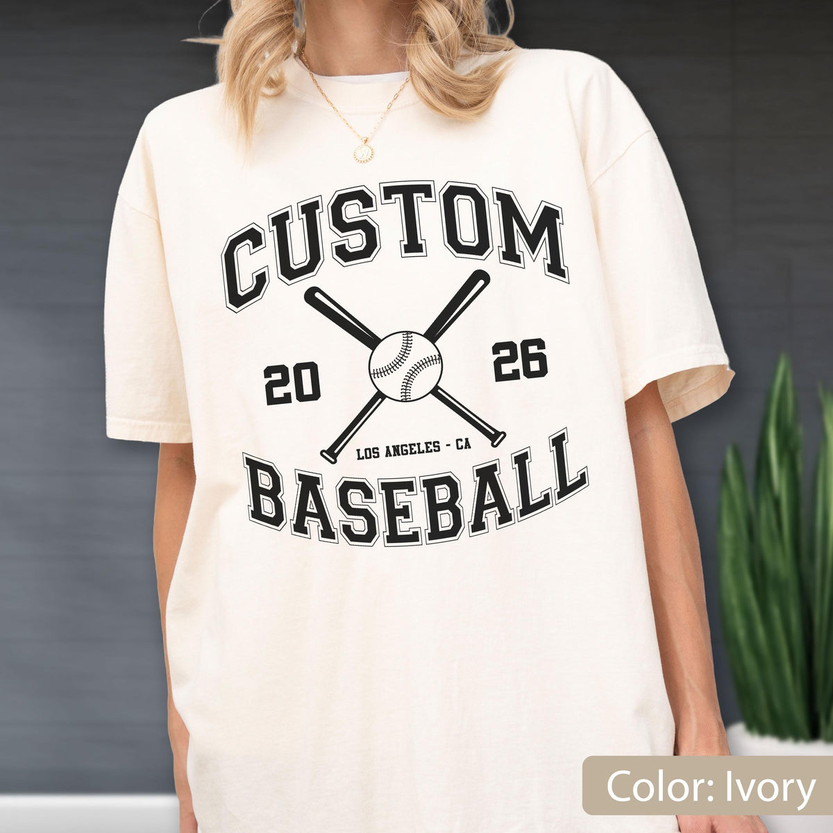 May include: An ivory short-sleeve t-shirt featuring a black graphic with the text CUSTOM BASEBALL, the numbers 20 and 26, and the location LOS ANGELES - CA. The design includes a central baseball and two crossed baseball bats.