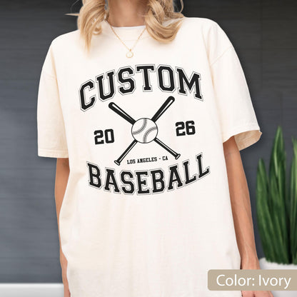 May include: An ivory short-sleeve t-shirt featuring a black graphic with the text CUSTOM BASEBALL, the numbers 20 and 26, and the location LOS ANGELES - CA. The design includes a central baseball and two crossed baseball bats.