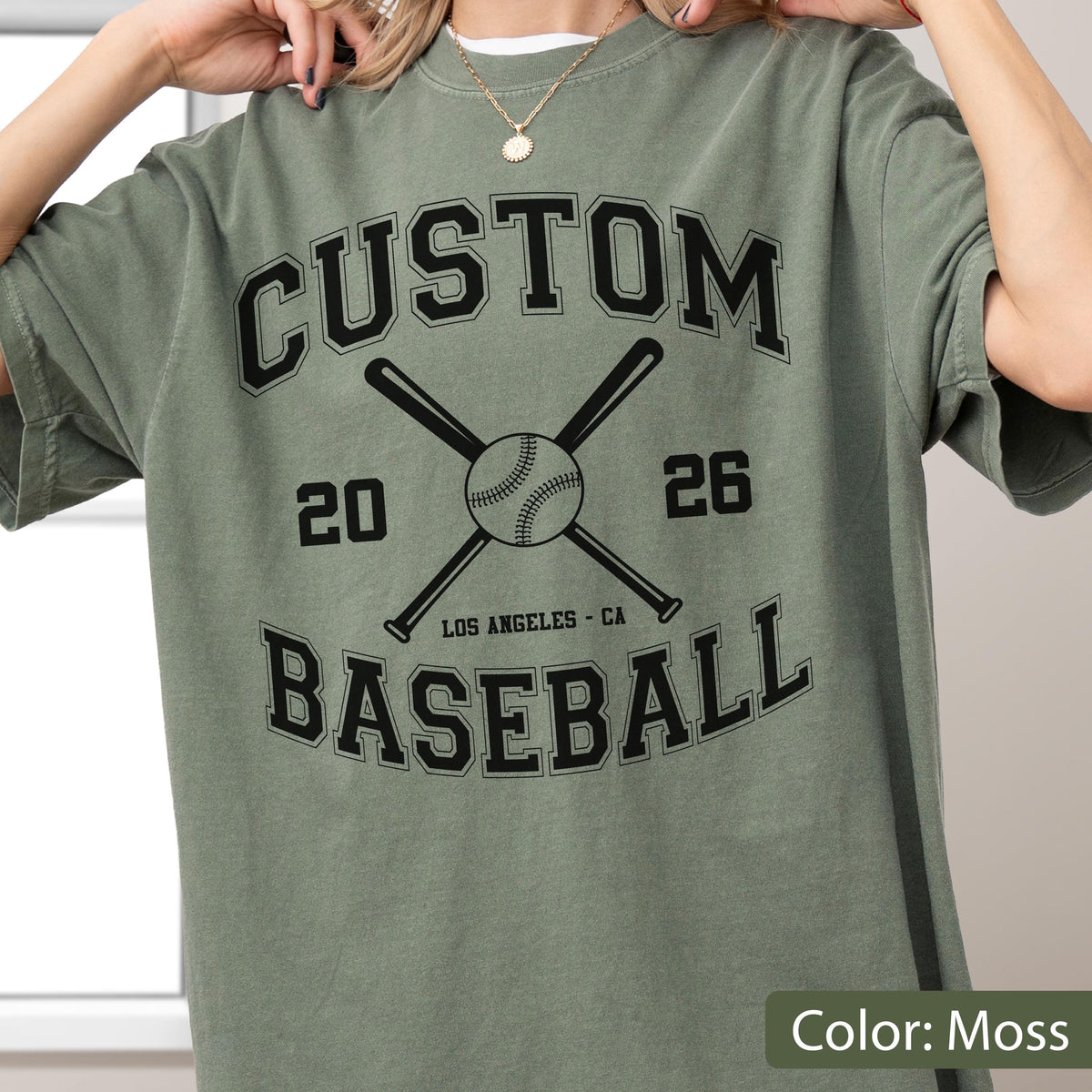 May include: A moss green short-sleeve t-shirt featuring a black graphic print. The design includes the text CUSTOM BASEBALL, the numbers 20 and 26, two crossed baseball bats, a baseball, and the text LOS ANGELES - CA in a collegiate font.
