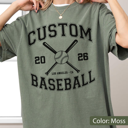 May include: A moss green short-sleeve t-shirt featuring a black graphic print. The design includes the text CUSTOM BASEBALL, the numbers 20 and 26, two crossed baseball bats, a baseball, and the text LOS ANGELES - CA in a collegiate font.