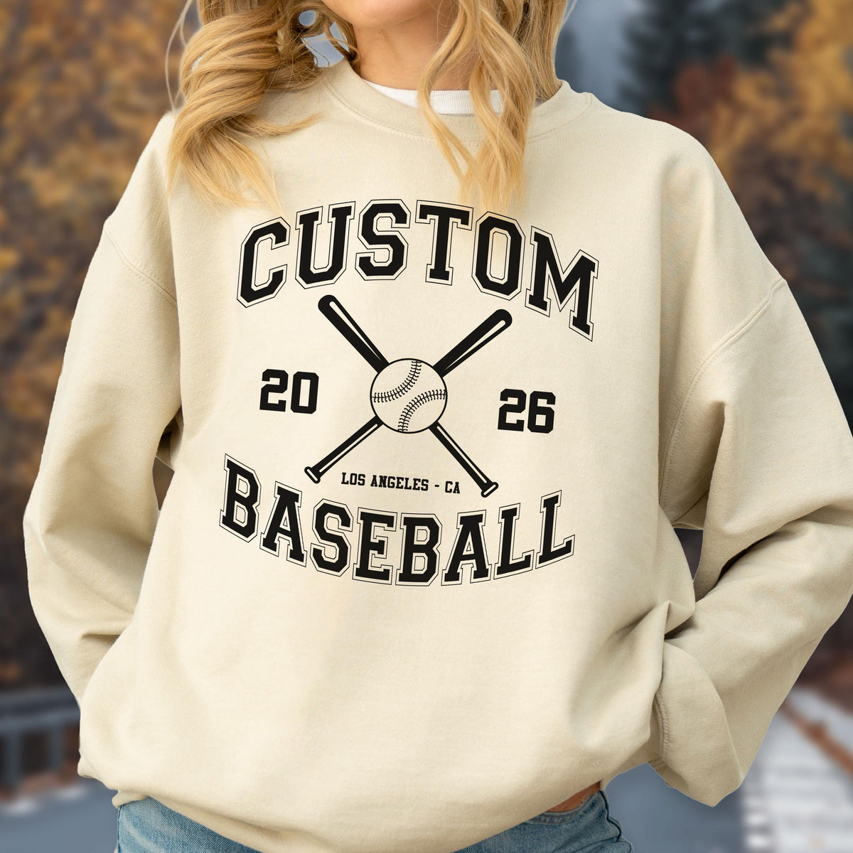 May include: A beige crewneck sweatshirt featuring a black graphic print with the text CUSTOM BASEBALL, the numbers 20 and 26, and the location LOS ANGELES - CA. The design includes a central baseball illustration with two crossed baseball bats.